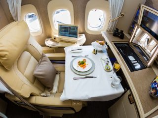 A luxurious airline suite with comfortable seats, table covered with white cloth with dinner plates, a laptop on the side and a large monitor at front