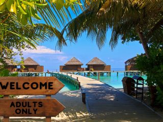 Photo showing a beach resort with huts and palm trees n the area with a wooden sign that reads Welcome, Adults only
